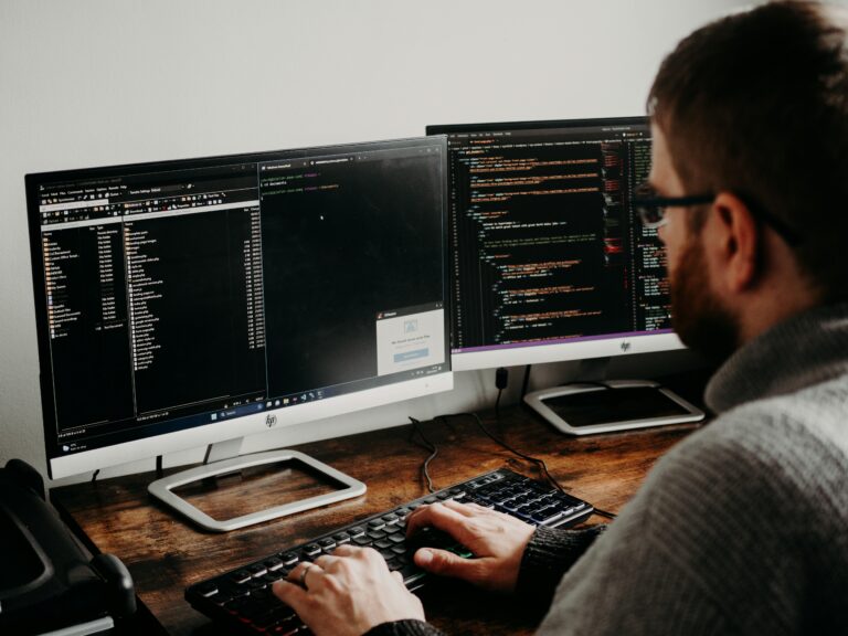 Person at home office desk with two monitors—one showing a file directory, the other code in an IDE. Face blurred; keyboard and office items visible.